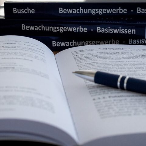 Aufgeschlagenes Buch mit einem Stift, im Hintergrund Bücher über Bewachungsgewerbe.