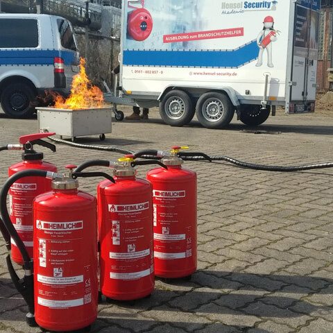 Rote Feuerlöscher stehen vor einem brennenden Behälter und einem Einsatzfahrzeug.